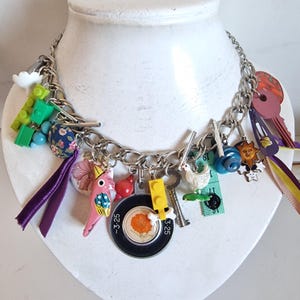 May include: A quirky necklace featuring a silver chain adorned with an assortment of colourful charms. Charms include a pink bird, keys, a toy block, a white cloud, and a black and orange dial. Purple and yellow ribbons hang from the necklace.