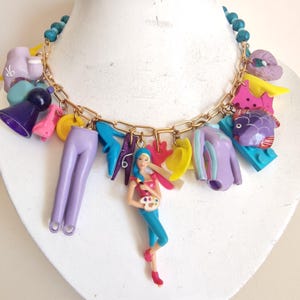 May include: A quirky necklace with a gold chain and a variety of colourful charms. The charms include miniature clothing, a doll figure, and other playful elements. The necklace has a turquoise bead strand.