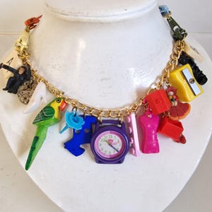 May include: A whimsical gold-tone charm necklace featuring a variety of colorful miniature charms. Charms include a parrot, high heel, watch, and other novelty items. The necklace is displayed on a white bust.