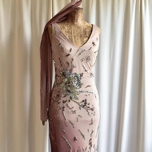 Könnte beinhalten: Ein bodenlanges, ärmelloses Kleid in einem Farbverlauf von Rosa zu Dunkelbraun. Das Kleid hat einen V-Ausschnitt und ist mit silbernen und grünen Pailletten in einem floralen Muster verziert. Ein passender Schal liegt über der Schulter der Büste.