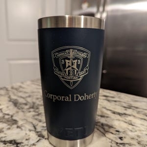 Puede incluir: Un vaso Yeti azul oscuro con tapa y base plateadas. Tiene un logotipo grabado en plata que dice "America's Battalion 3rd 3rd Fortes Fortuna Juvat" y el nombre "Corporal Doherty".
