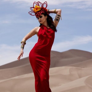 May include: A woman in a red dress and a floral headpiece poses in a desert landscape. The dress is long and flowing, and the headpiece is made of red and yellow flowers.