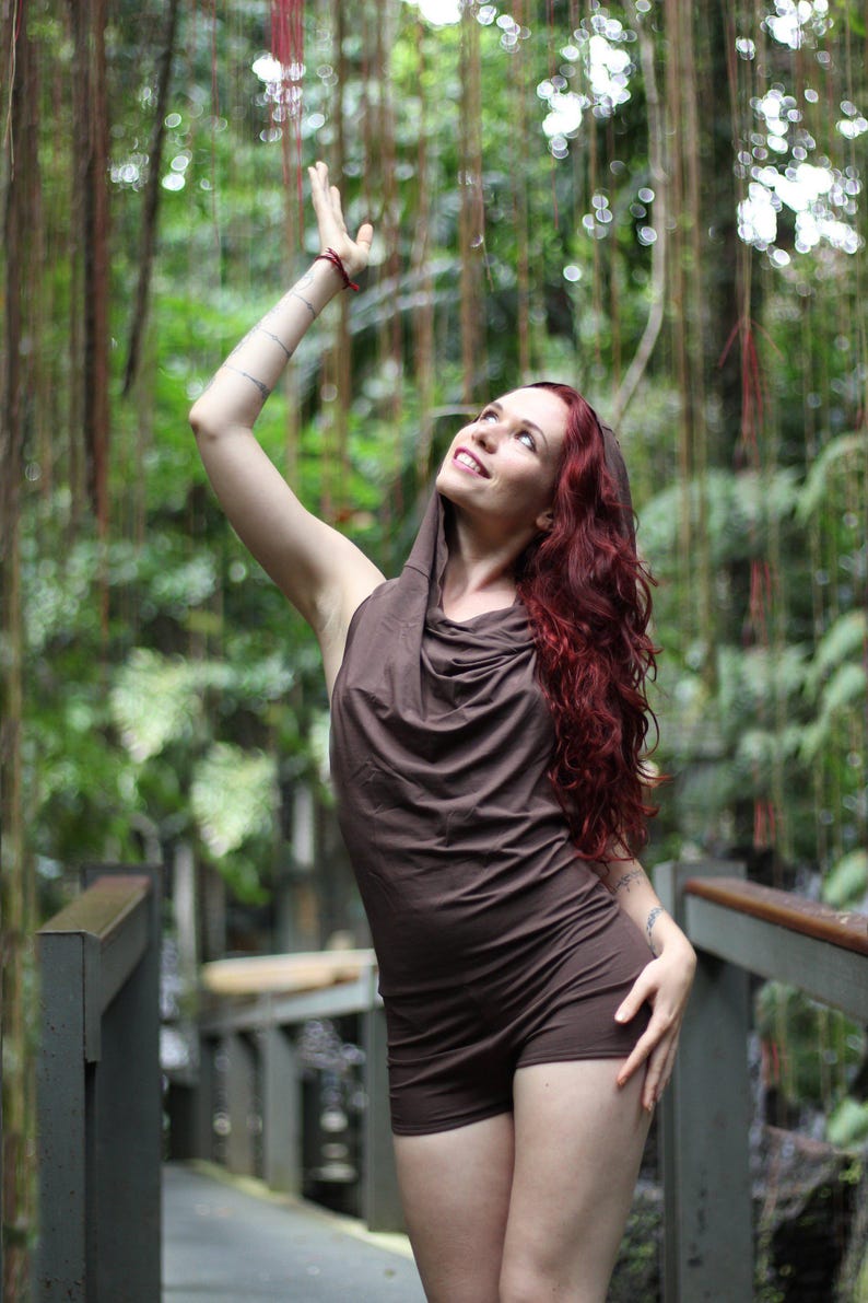May include: A woman wearing a brown hooded jumpsuit with a cowl neck, standing in a lush green forest setting. She is looking up and has her arm raised.