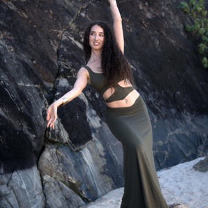 Puede incluir: Una mujer con un atuendo verde ajustado con recortes en la cintura, posa sobre una roca con el brazo levantado. Tiene el pelo largo, oscuro y rizado y lleva un piercing en el ombligo plateado.