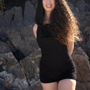 May include: A woman with long curly hair is wearing a black hooded top and shorts. She is standing in front of a rocky background.