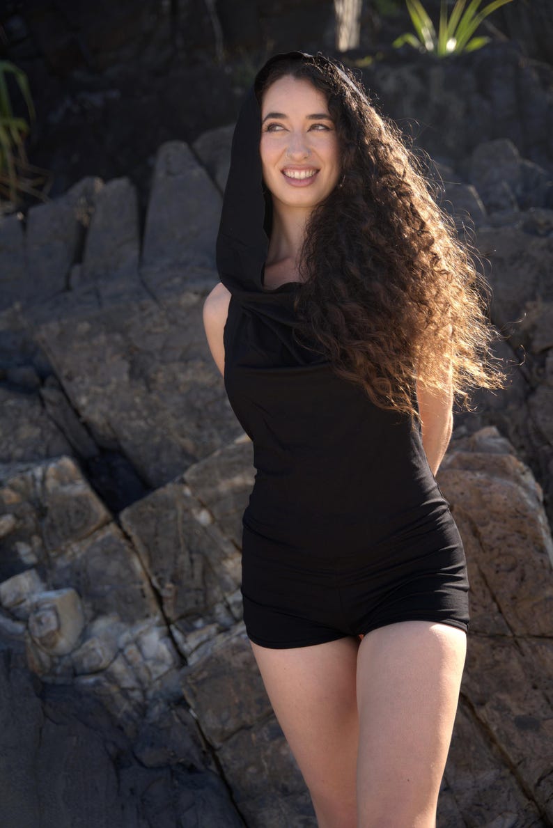 May include: A woman with long curly hair is wearing a black hooded top and shorts. She is standing in front of a rocky background.