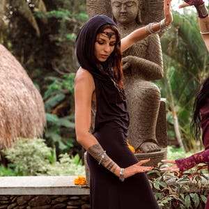 May include: A woman wearing a black hooded dress and silver bracelets, posing in front of a stone Buddha statue. The dress is sleeveless and flows to the floor. The woman is wearing a jeweled headpiece. The background includes lush greenery and stone walls.