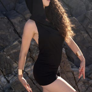 May include: A woman wearing a black hooded dress with a cowl neck. She is standing in front of a rocky background.