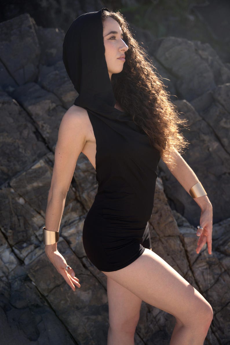 May include: A woman wearing a black hooded dress with a cowl neck. She is standing in front of a rocky background.