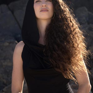 May include: A woman with long, curly brown hair wears a black sleeveless top with a cowl neck and a hood. She is wearing a gold bracelet on her left wrist.