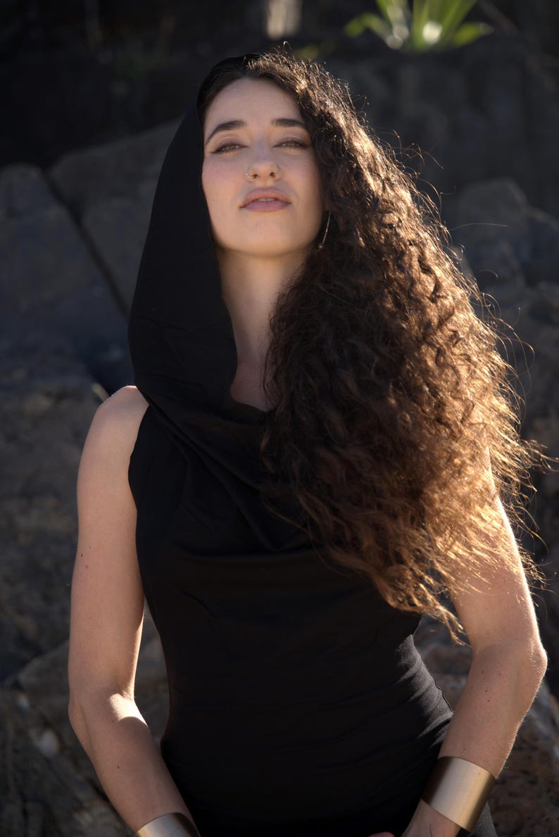 May include: A woman with long, curly brown hair wears a black sleeveless top with a cowl neck and a hood. She is wearing a gold bracelet on her left wrist.