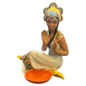 May include: A ceramic figurine of a seated woman with dark hair and a yellow and blue headdress. She wears a white dress and has her hands raised. An orange bowl sits in front of her. The figurine has colourful accents and is likely a decorative item.