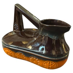 Puede incluir: Una jarra de cerámica marrón con un asa única y un diseño decorativo naranja y marrón. La jarra tiene un cuerpo redondeado y un pico ancho.