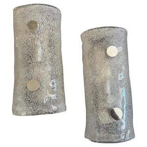 May include: Two textured, cylindrical glass sconces with a silver-gray hue. Each sconce features two circular, silver-colored accents. The glass has a speckled appearance, and the sconces have a slightly flared base and top.