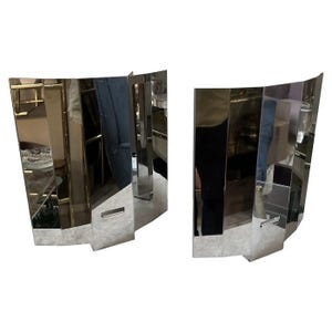 May include: Two mirrored wall sconces with a geometric design. The sconces are made of multiple rectangular and trapezoidal mirrored pieces, creating a reflective surface. The sconces have a curved shape and are designed to be mounted on a wall.