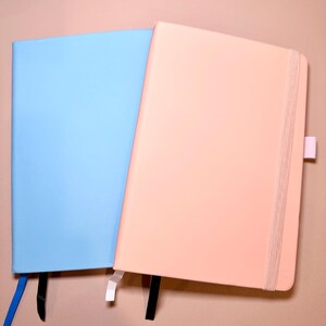 Puede incluir: Dos cuadernos, uno azul claro y otro rosa claro, con marcadores de cinta negra y blanca. Los cuadernos están cerrados y colocados planos sobre una superficie rosa claro.