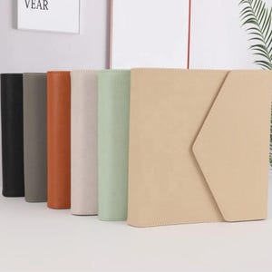 May include: Six different coloured leather-bound journals with a flap closure. The journals are arranged in a row from left to right, with the black journal on the left and the beige journal on the right.