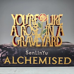 May include: A gold and red decorative sign with the phrase "YOU'RE LIKE A ROSE IN A GRAVEYARD" and a rose illustration. The sign is placed on top of a book with the title "ALCHEMISED".