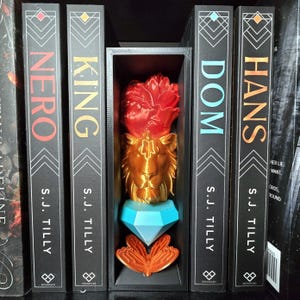 May include: A black rectangular display box containing a stacked sculpture of a red flower, a gold lion head, a light blue diamond shape, and an orange butterfly. The box is positioned between four books titled NERO, KING, DOM, and HANS by S.J. Tilly.