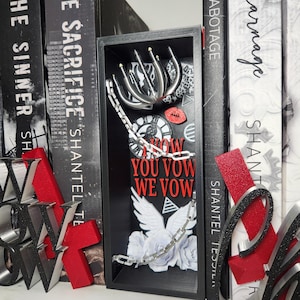 May include: A black shadow box with a variety of decorative elements. It features a silver chain, a white dove, a red lip, and the words "NOW YOU VOW WE VOW" in red. The background includes a leopard print and clock faces.