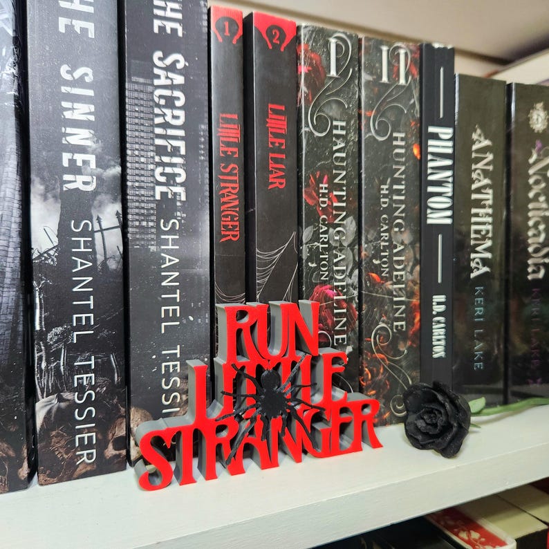 Puede incluir: Una estanter&iacute;a con varios libros de cubiertas oscuras. Un letrero rojo y gris impreso en 3D que dice "Run Little Stranger" est&aacute; frente a los libros. Una rosa negra est&aacute; en el estante.