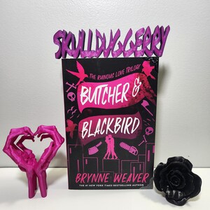 May include: A book cover for "Butcher & Blackbird" by Brynne Weaver, with a black background and pink text. The cover features a pink heart-shaped sculpture, a black rose, and a purple "SKULLDUGGERY" book stand.