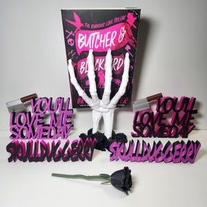 May include: A display featuring a book titled "Butcher & Blackbird" with a skeleton hand, text that reads "You'll Love Me Someday" and "Skullloveberry", a cleaver, and a black rose. The book and text are in black and pink.