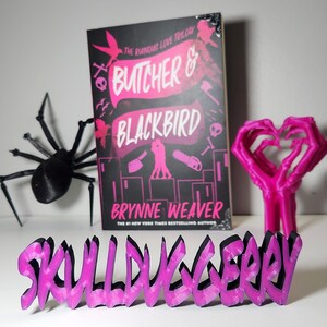 May include: A book cover for "Butcher & Blackbird" by Brynne Weaver, with a black background and pink text. A black spider, a pink heart-shaped hand sculpture, and a pink and black "Skull Loveberry" sign are also present.