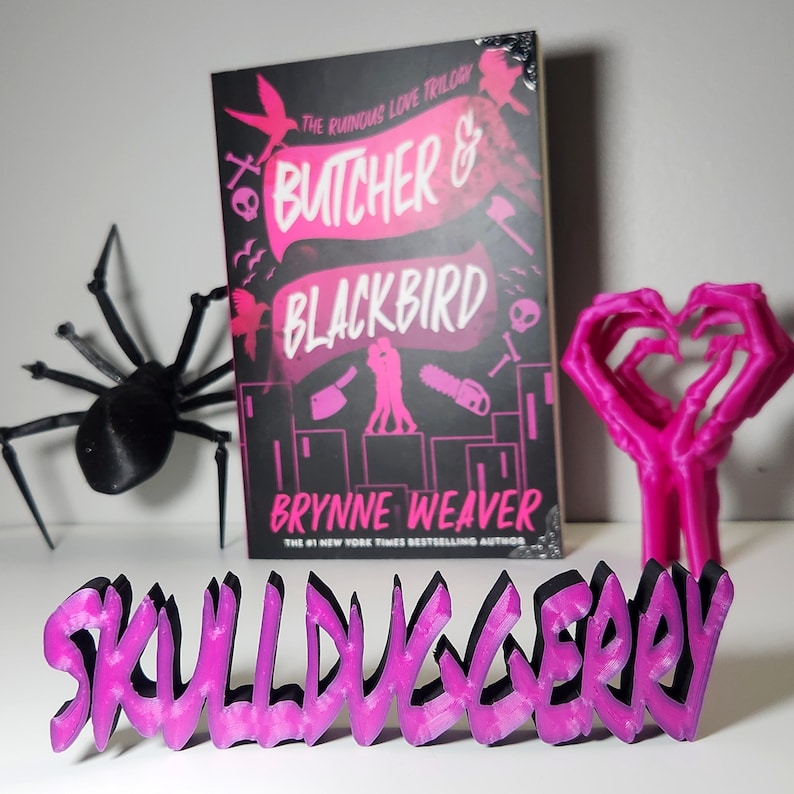 May include: A book cover for "Butcher & Blackbird" by Brynne Weaver, with a black background and pink text. A black spider, a pink heart-shaped hand sculpture, and a pink and black "Skull Loveberry" sign are also present.