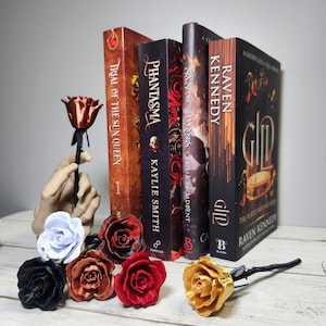 May include: A display of fantasy novels and decorative metal roses in various colors. The books include titles like "Trial of the Sun Queen" and "Raven Kennedy." The roses are in black, red, gold, and other hues.