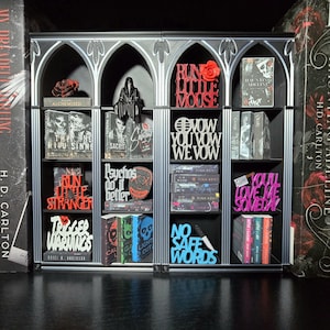 May include: A miniature gothic bookshelf display featuring four vertical sections filled with small book replicas and decorative signs. The shelves contain various items with text including Run Little Mouse, You Vow We Vow, Psychos Do It Better, Trigger Warnings, No Safe Words, and You'll Love Me Someday, alongside miniature figures and books.