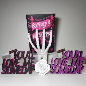 May include: A book titled "Butcher & Blackbird" with a white skeleton hand, two decorative signs that read "You'll Love Me Someday", a white rose, and two cleavers with brown handles.
