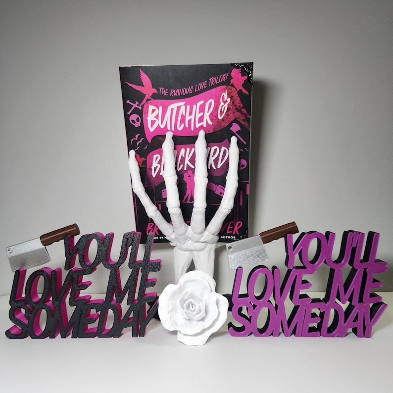 May include: A book titled "Butcher & Blackbird" with a white skeleton hand, two decorative signs that read "You'll Love Me Someday", a white rose, and two cleavers with brown handles.