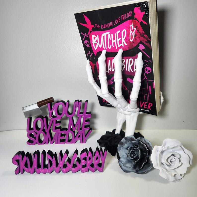 May include: A white skeleton hand book holder supporting a book titled "Butcher & Blackbird". The image also includes a pink and black "You'll Love Me Someday" sign, a "Skullberry" sign, a cleaver, and two rose sculptures.