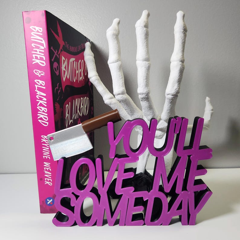 May include: A book titled "Butcher & Blackbird" with a pink cover, a white skeleton hand, a cleaver, and a purple sign that reads "You'll Love Me Someday". The book and sign are on a white surface.