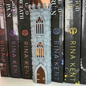 May include: A gray, textured, 3D-printed castle tower featuring a brown arched door with gold accents and two arched windows with vertical bars. The structure has a crenelated top and emits a warm, internal orange glow through the windows.