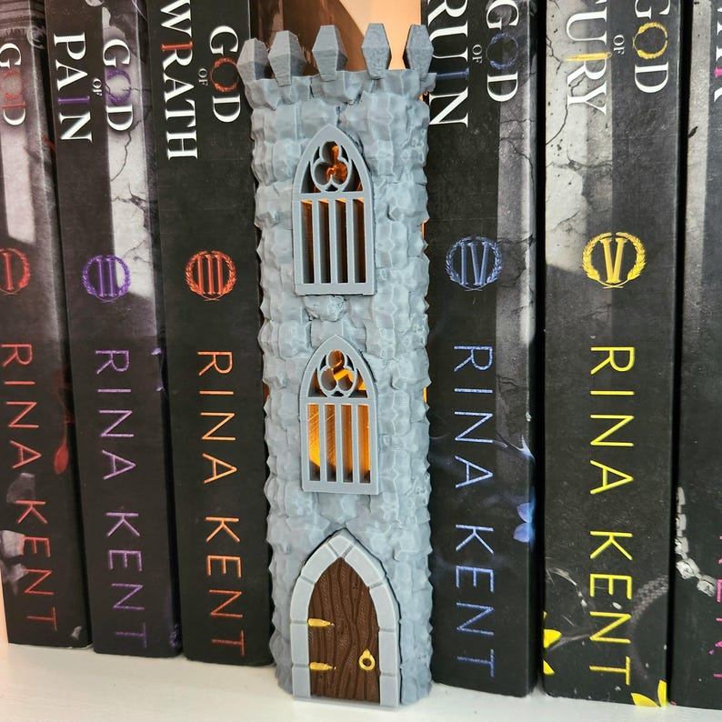May include: A gray, textured, 3D-printed castle tower featuring a brown arched door with gold accents and two arched windows with vertical bars. The structure has a crenelated top and emits a warm, internal orange glow through the windows.