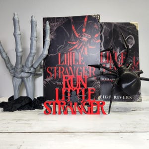 Puede incluir: Una exhibici&oacute;n tem&aacute;tica de Halloween con un libro titulado "Little Stranger Run Little Stranger" con una portada roja y negra. Una ara&ntilde;a negra y un letrero rojo con el t&iacute;tulo del libro est&aacute;n frente al libro. Una mano de esqueleto gris est&aacute; a la izquierda.