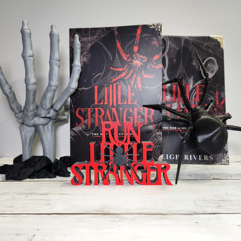 Puede incluir: Una exhibici&oacute;n tem&aacute;tica de Halloween con un libro titulado "Little Stranger Run Little Stranger" con una portada roja y negra. Una ara&ntilde;a negra y un letrero rojo con el t&iacute;tulo del libro est&aacute;n frente al libro. Una mano de esqueleto gris est&aacute; a la izquierda.