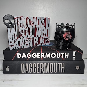 May include: A black skull with a silver crown and a pink rose on its cheek, next to a silver sign with red accents reading THE CRACKS IN MY SOUL ARENT BROKEN PLACE, all resting on two stacked books titled DAGGERMOUTH by H.M. Wolfe.