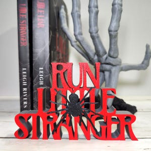 Puede incluir: Decoraci&oacute;n de Halloween roja y negra con la frase "RUN LITTLE STRANGER" y una ara&ntilde;a negra con purpurina. Dos libros est&aacute;n detr&aacute;s de la decoraci&oacute;n y una mano de esqueleto gris en el fondo.