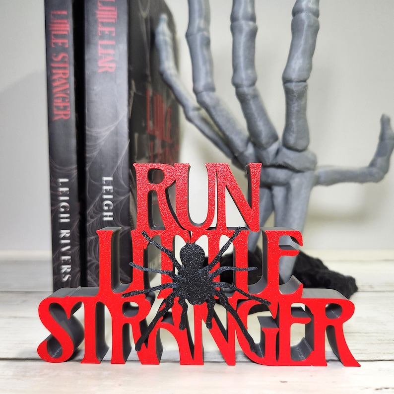 Puede incluir: Decoraci&oacute;n de Halloween roja y negra con la frase "RUN LITTLE STRANGER" y una ara&ntilde;a negra con purpurina. Dos libros est&aacute;n detr&aacute;s de la decoraci&oacute;n y una mano de esqueleto gris en el fondo.