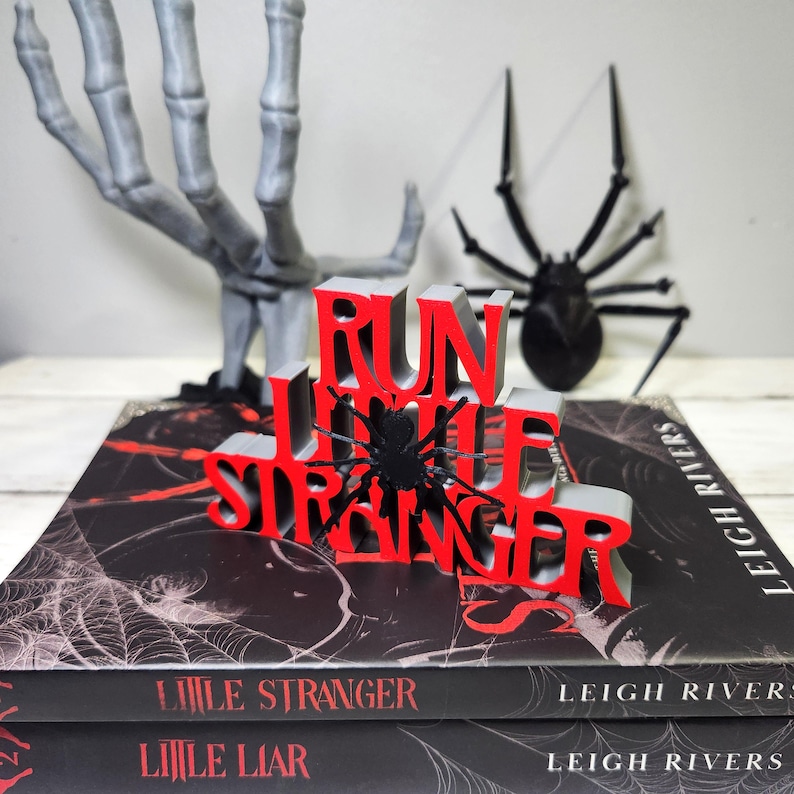 Puede incluir: Una decoraci&oacute;n tem&aacute;tica de Halloween con las palabras "RUN LITTLE STRANGER" en rojo y gris, con una ara&ntilde;a negra. Descansa sobre una pila de libros titulados "LITTLE STRANGER" y "LITTLE LIAR". Una mano de esqueleto gris y una ara&ntilde;a negra est&aacute;n en el fondo.