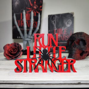 Puede incluir: Un letrero de madera rojo y negro con las palabras "RUN LITTLE STRANGER" y una ara&ntilde;a negra. El letrero se muestra con una rosa roja, una calavera y dos libros en el fondo.