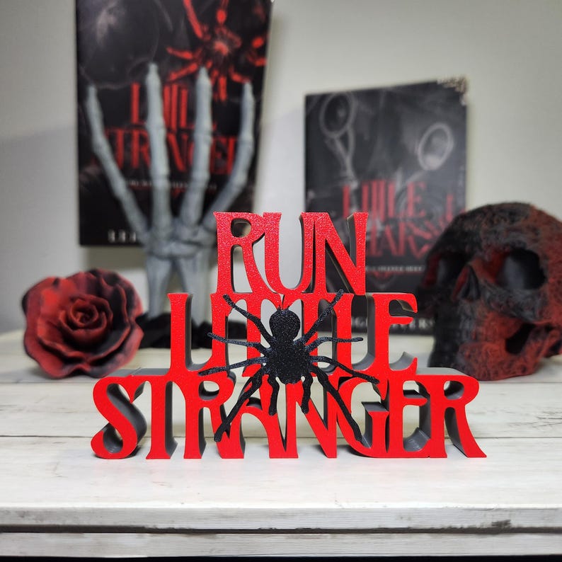 Puede incluir: Un letrero de madera rojo y negro con las palabras "RUN LITTLE STRANGER" y una ara&ntilde;a negra. El letrero se muestra con una rosa roja, una calavera y dos libros en el fondo.