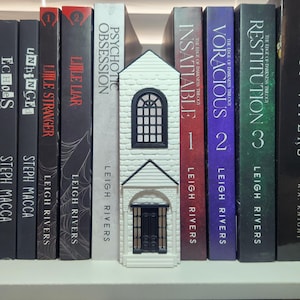 May include: A white, three-dimensional, house-shaped bookend with a black arched window and a black door, positioned between several books on a shelf. The bookend features a textured brick-like facade and a peaked roof design.