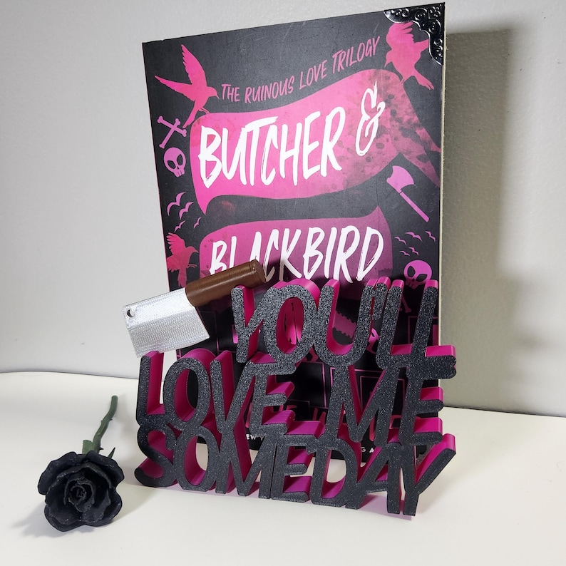 May include: A book cover for "Butcher & Blackbird" with pink and black text and graphics. A decorative sign reads "You'll Love Me Someday" in black and pink. A small cleaver and a black rose are also present.