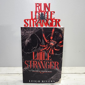 Puede incluir: Un libro titulado "Little Stranger" con un gr&aacute;fico de ara&ntilde;a roja y negra. El libro est&aacute; coronado con un letrero rojo que dice "Run Little Stranger" con una ara&ntilde;a negra. La portada del libro presenta un dise&ntilde;o de telara&ntilde;a.
