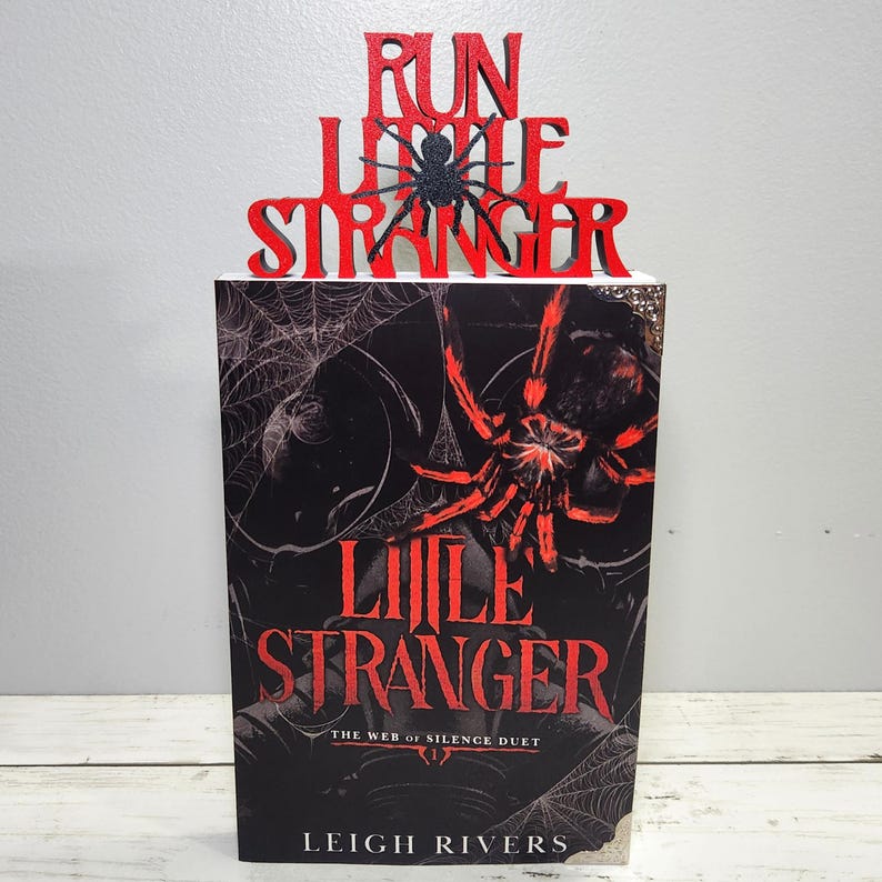Puede incluir: Un libro titulado "Little Stranger" con un gr&aacute;fico de ara&ntilde;a roja y negra. El libro est&aacute; coronado con un letrero rojo que dice "Run Little Stranger" con una ara&ntilde;a negra. La portada del libro presenta un dise&ntilde;o de telara&ntilde;a.