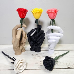 May include: A collection of rose-holding hand sculptures in beige, black, and white, each holding a rose in red, yellow, and pink. Two additional roses, one white and one black, are displayed separately. The roses have green stems.